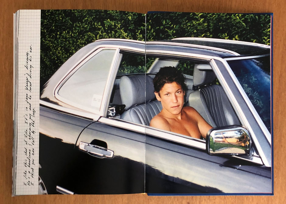 A woman with short dark hair sitting topless inside a classic black car with a gray interior, parked outdoors with lush green foliage in the background.
