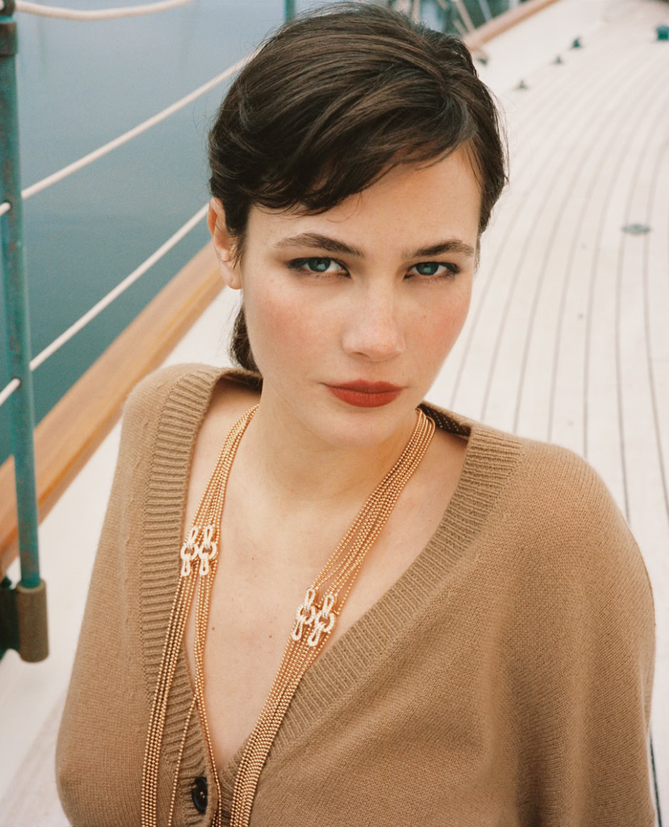 Elegant portrait of a woman with short dark hair and blue eyes, wearing a beige sweater and layered gold jewelry, on a boat deck with water in the background.