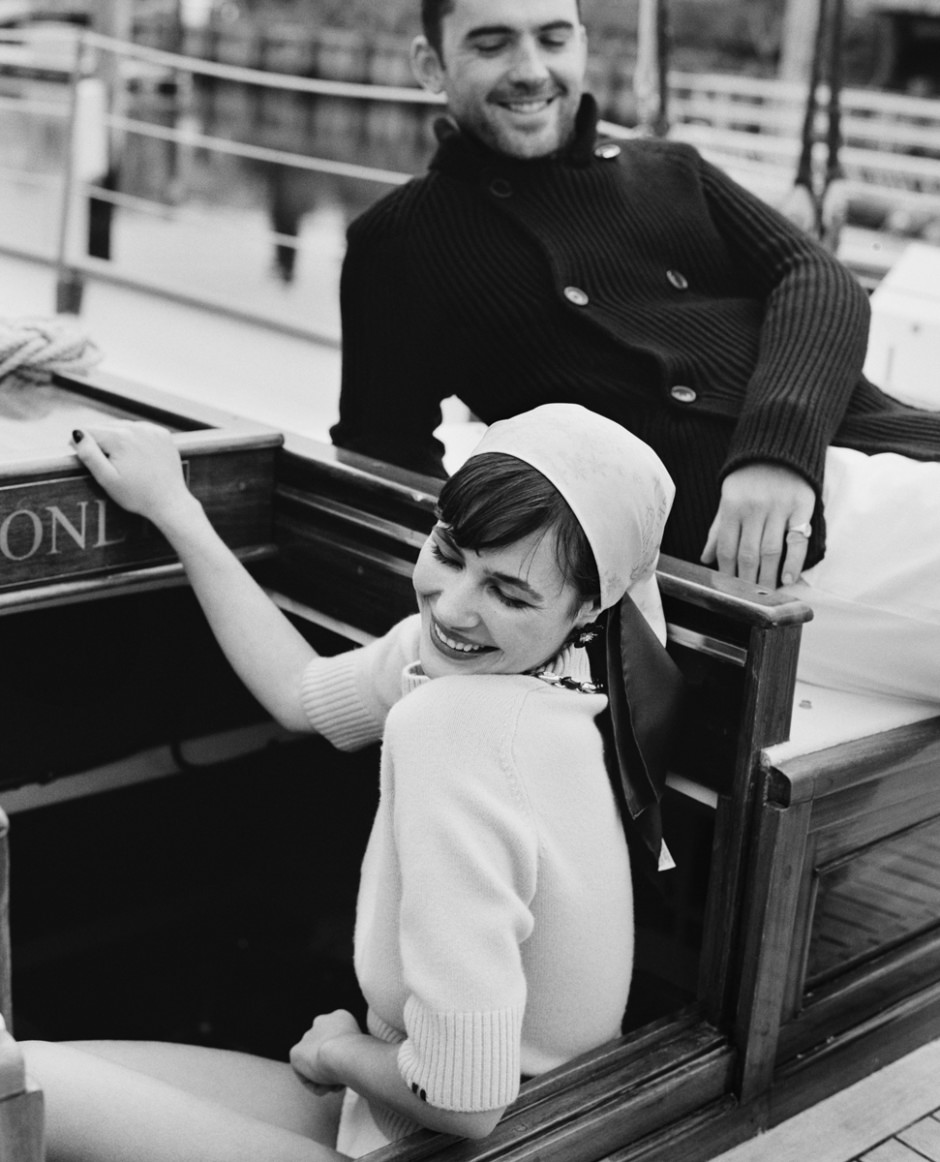 A woman laughing on a boat, wearing a headscarf and sweater, with a man smiling behind her, captured in black and white, showcasing candid portrait photography, joyful moments, and lifestyle photography.